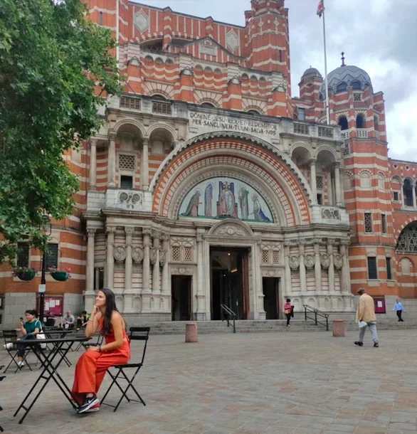 Vista exterior de la Catedral de Westminster, destacando su impresionante fachada de ladrillo rojo y detalles arquitectónicos, con una persona sentada en una mesa de café en primer plano