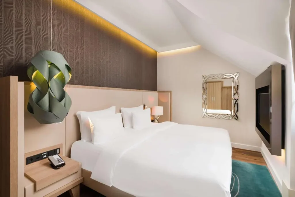 Habitación de hotel luminosa y moderna en Budapest, con cama grande de sábanas blancas, lámpara de diseño original, espejo decorativo, televisión empotrada y detalles en madera y tonos neutros que crean un ambiente elegante y acogedor