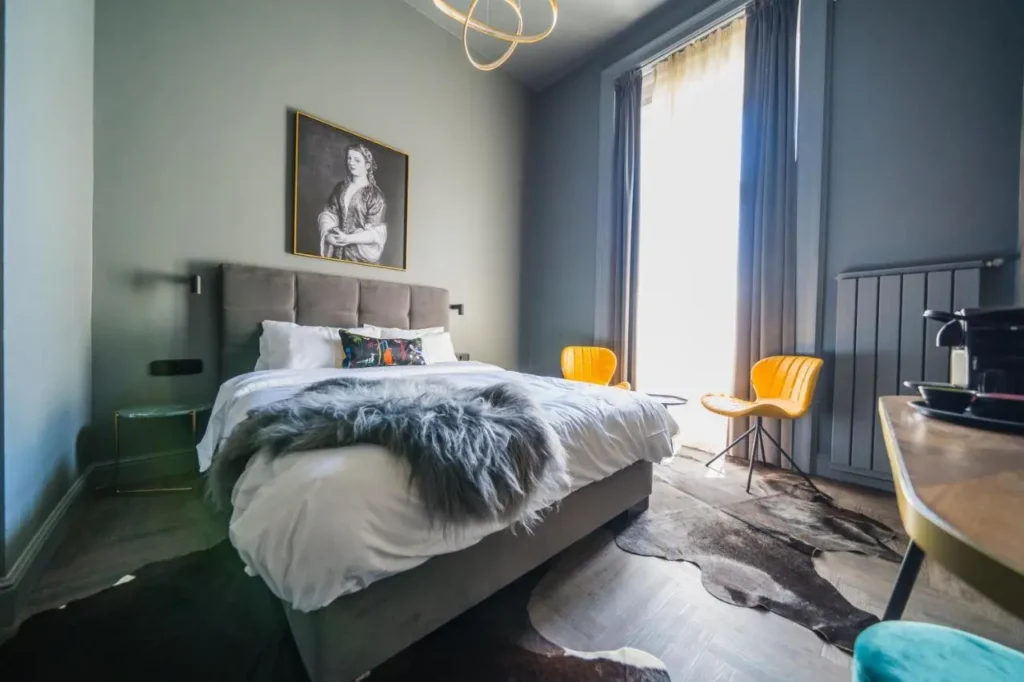 Habitación moderna y elegante en un hotel boutique de Budapest, con paredes en tonos oscuros, una cama grande con textiles de lujo, sillas amarillas de diseño, una lámpara colgante sofisticada y una gran ventana que deja entrar luz natural.