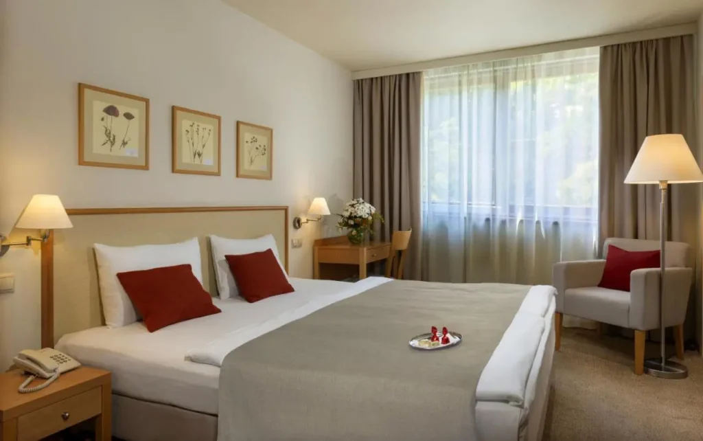 Habitación de hotel luminosa y moderna con cama de matrimonio, ropa de cama blanca y detalles en rojo. La habitación cuenta con un gran ventanal con cortinas claras, un sillón con cojín rojo a juego, un escritorio de madera con una silla, y dos lámparas de noche a cada lado de la cama.