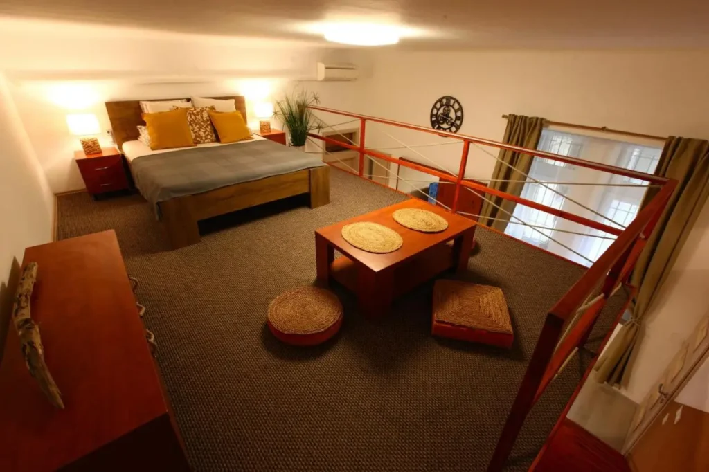 Habitación tipo loft en Budapest con diseño acogedor y rústico, cama de madera con cojines en tonos cálidos, iluminación tenue, mesa baja de estilo japonés con cojines de suelo y vista a la planta baja a través de una barandilla roja.