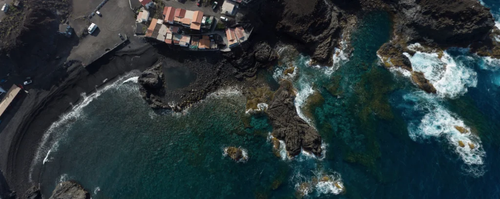 Vista aérea de la Playa de Punta Larga, en La Palma, Canarias. La imagen muestra el contraste de la costa volcánica, con agua cristalina y rocas dispersas en el mar. A lo lejos, se observan varias casas en las cercanías de la playa