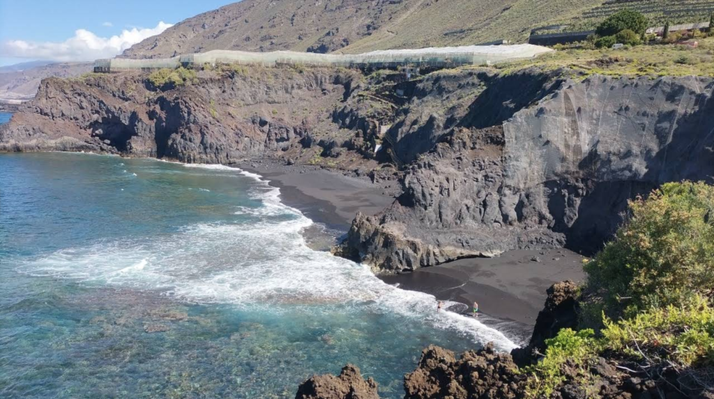 Playa de La Zamora, en La Palma, Canarias, mostrando un paisaje impresionante de acantilados volcánicos que caen hacia el mar. La arena negra característica de la isla contrasta con el agua cristalina. Se puede ver a algunas personas caminando por la orilla, disfrutando de este entorno natural único