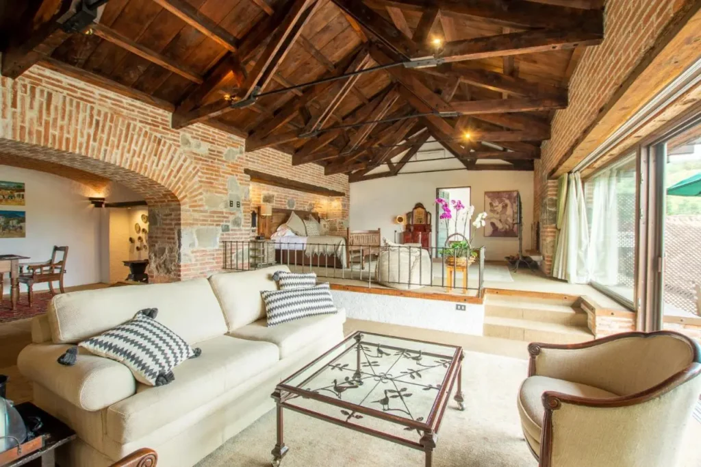 Interiores de una habitación de hotel de lujo Antigua Guatemala con techo de vigas de madera expuestas, ladrillos expuestos en las paredes y una decoración elegante con muebles clásicos. Espacio amplio y luminoso con sala de estar, mesa de comedor, plantas decorativas y ventanales que ofrecen vistas panorámicas