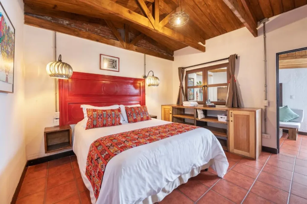 Habitación de hotel en Antigua Guatemala con cama de matrimonio y cabecera roja decorada con textiles tradicionales. Techo de madera expuesta, lámparas colgantes elegantes y suelo de cerámica terracota. Ambiente acogedor y moderno con muebles de madera y decoración minimalista