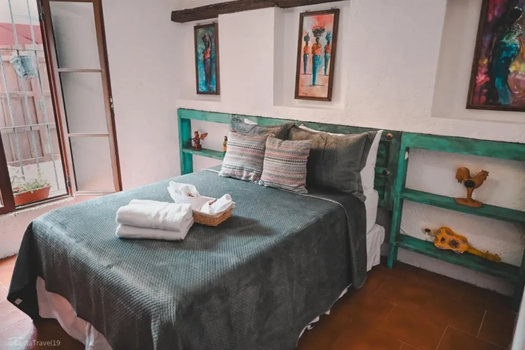 Habitación acogedora en Antigua Guatemala con cama de matrimonio, cabecera decorada con madera verde y adornos artesanales. La habitación cuenta con cuadros de arte local, sábanas y almohadas en tonos grises, y una ventana con vistas al patio. Detalles de decoración tradicional, como una guitarra y una figura de gallo en las estanterías