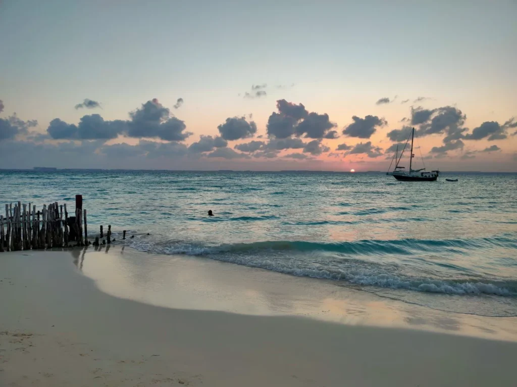 Atardecer en Playa Centro de Isla Mujeres con velero en el horizonte y cielo rosado