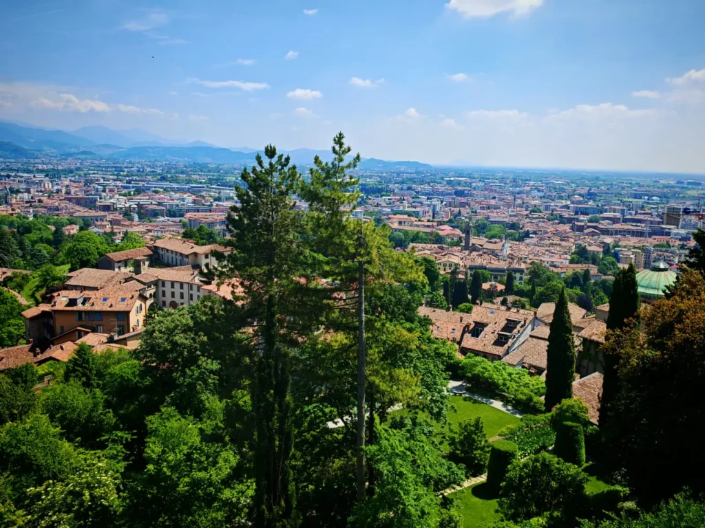 Vista panorámica de Bérgamo desde la Città Alta con tejados medievales, vegetación y la llanura lombarda al fondo.