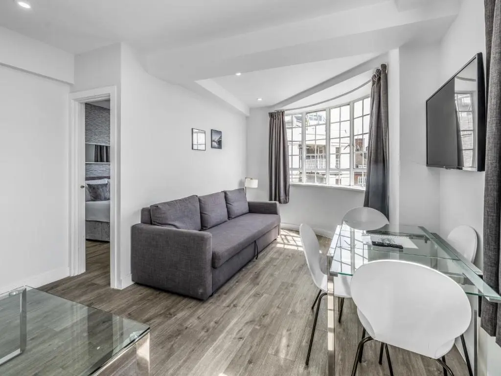 Apartamento donde alojarse en Londres con salón luminoso, sofá gris, ventanal curvo y mesa de comedor de cristal; opción de alojamiento céntrico ideal para familias o parejas.