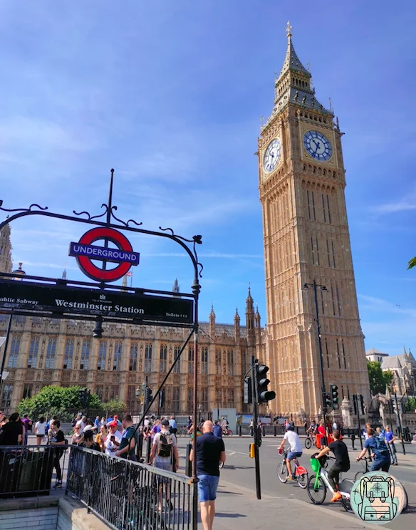 Vista del Big Ben y la estación de metro Westminster en Londres, con el icónico cartel de Underground y turistas caminando y montando bicicletas en una soleada mañana