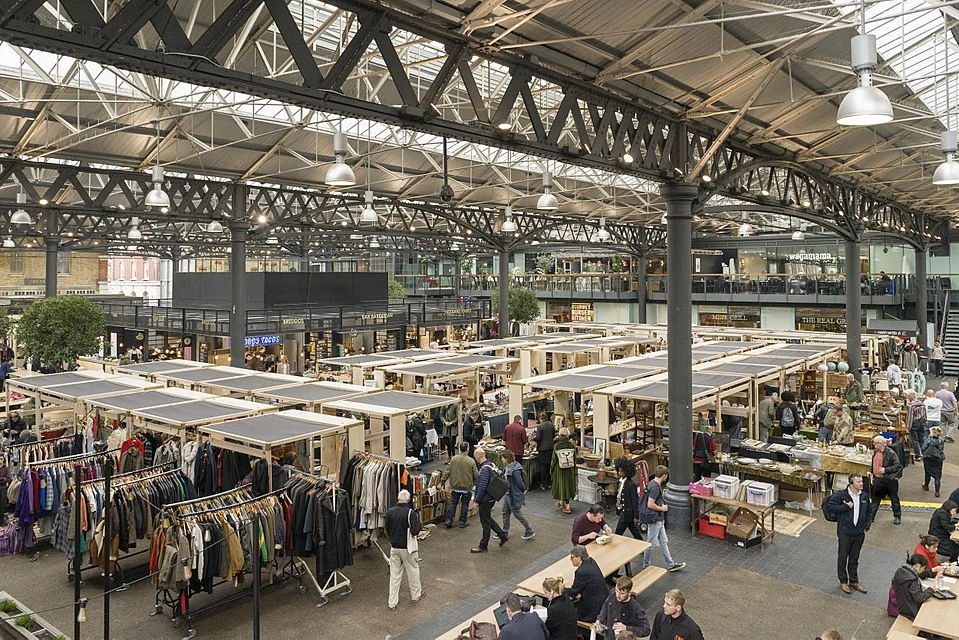 L4.22 Vista panorámica del interior del mercado de Spitalfields, con puestos de ropa vintage, artículos de segunda mano y puestos de comida, todo bajo una estructura de hierro industrial
