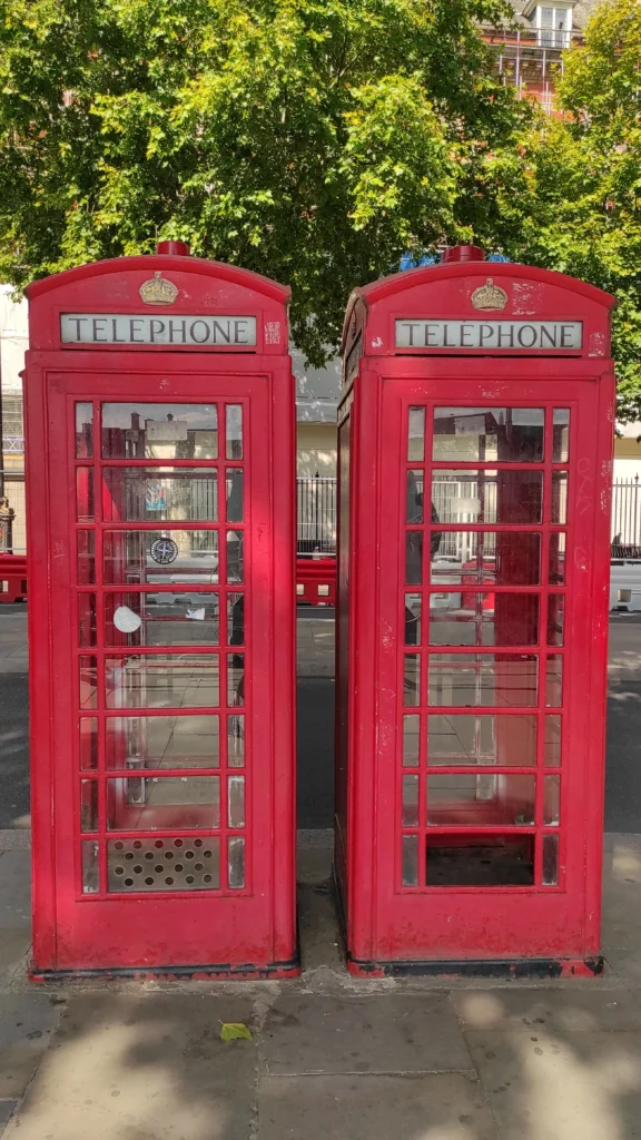 L4.22 Dos cabinas telefónicas rojas clásicas de Londres, etiquetadas como 'TELEPHONE', ubicadas en una calle arbolada con un ambiente soleado y tranquilo