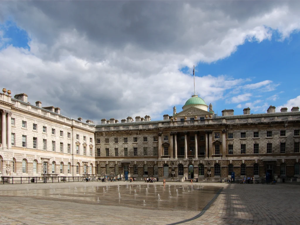 Vista exterior del Somerset House en Londres, con su elegante arquitectura neoclásica, una plaza central con fuentes de agua y un cielo parcialmente nublado