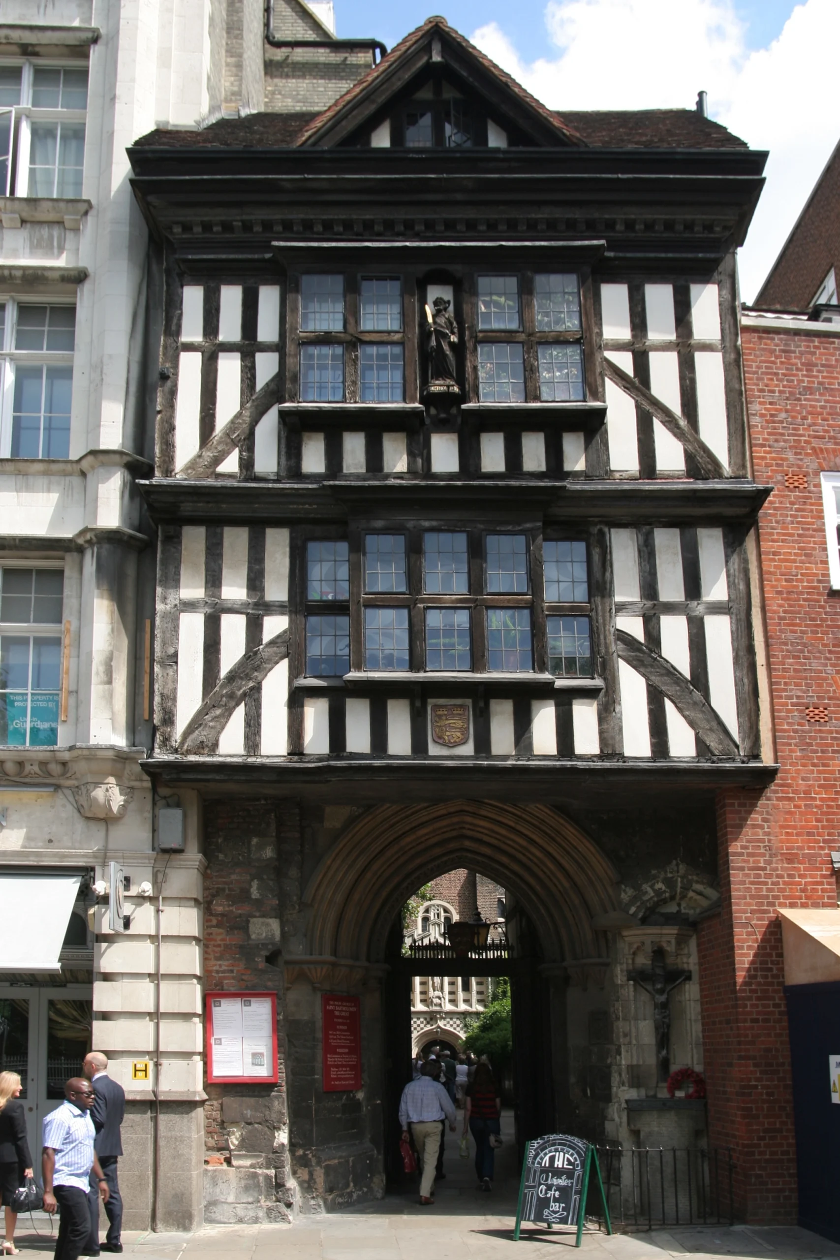 L4.22 Vista del St. Bartolomew Gatehouse, una estructura medieval de madera y piedra en el corazón de Londres, con una figura en la parte superior y un arco de acceso que lleva a un patio histórico