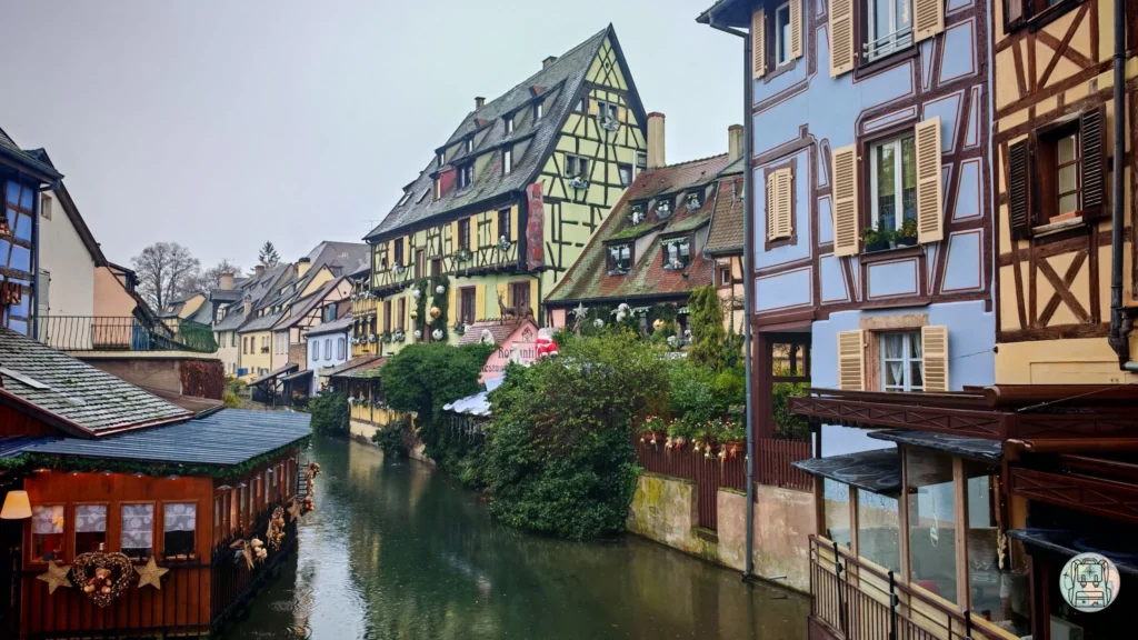 Vista de un canal en el barrio de la 'Petite Venise' en Colmar, Francia, con casas tradicionales de entramado de madera de colores pasteles decoradas con adornos navideños, reflejándose en el agua bajo un cielo invernal