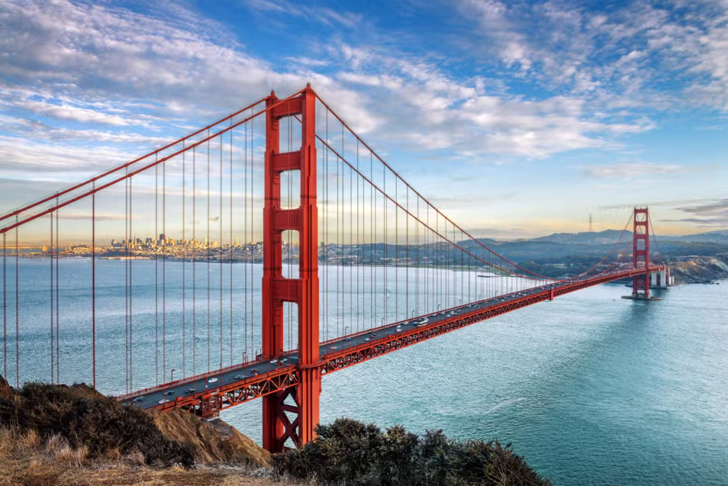 Vista panorámica del Puente Golden Gate en San Francisco desde un mirador elevado, mostrando su característica estructura de acero naranja rojizo cruzando la bahía bajo un cielo azul con nubes ligeras al atardecer. Donde viajar en agosto