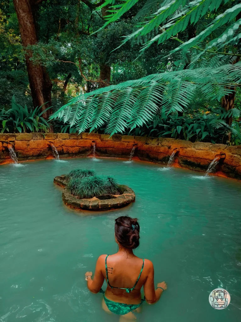Vista desde atrás de una mujer con bañador verde sumergida en una piscina de aguas termales de color turquesa en el Parque Terra Nostra, Azores. El agua brota de varios caños en un muro de piedra circular rodeado de frondosa vegetación tropical y helechos gigantes. Azores es un destino ideal para visitar en septiembre