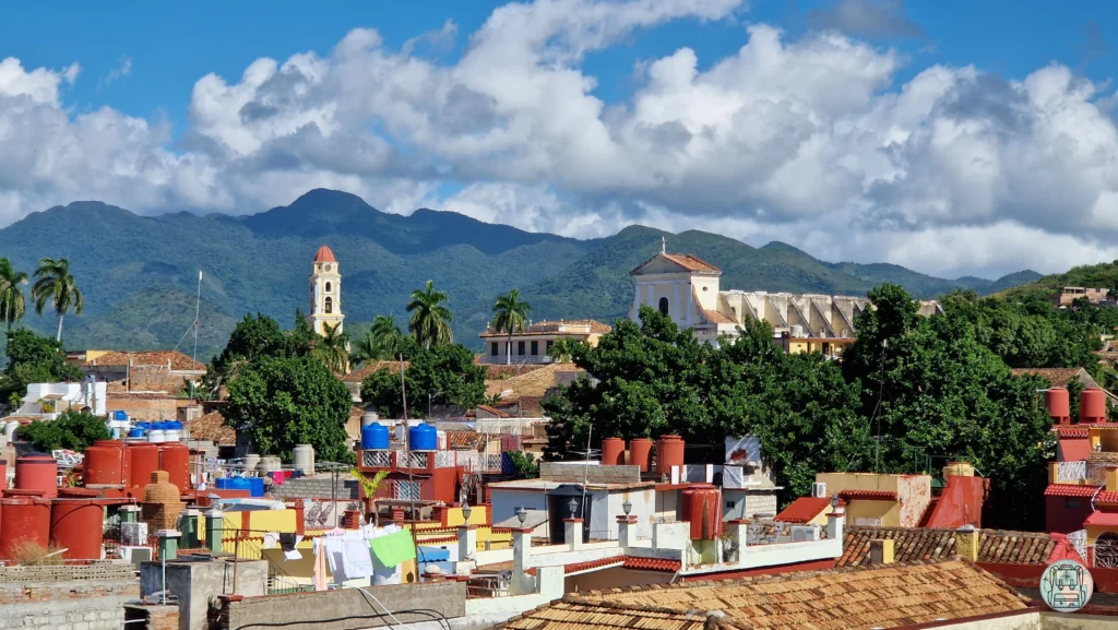 Vista de los tejados coloniales de Trinidad, Cuba, con la torre amarilla del campanario de San Francisco de Asís, palmeras y las montañas de la Sierra del Escambray bajo un cielo con nubes blancas. Donde viajar en enero