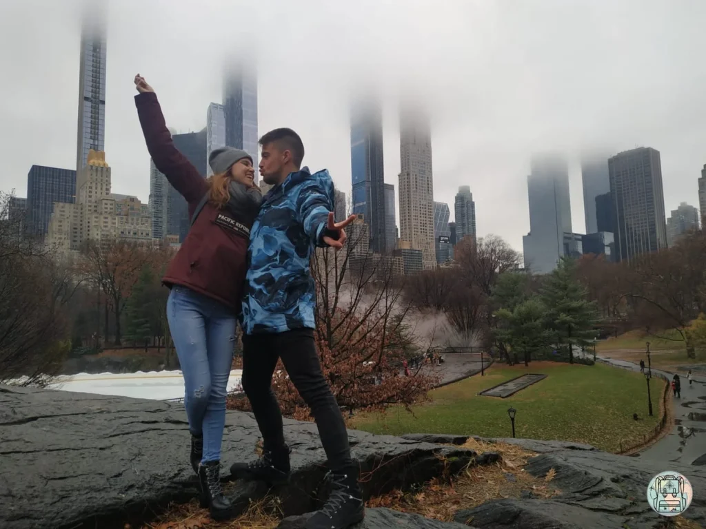 Una pareja joven posa de forma divertida sobre una roca en Central Park, Nueva York. Se ve al chico lanzando un beso y a la chica con el brazo en alto bajo un cielo nublado y neblinoso que envuelve los rascacielos del Midtown al fondo. Donde viajar en diciembre