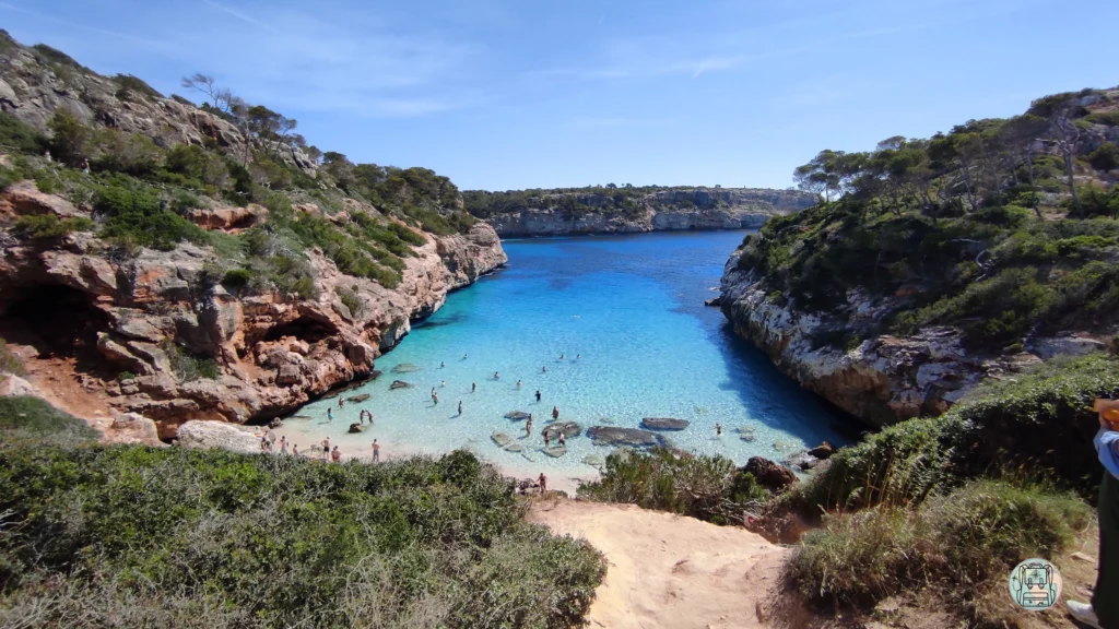 Caló des Moro en Mallorca, una pequeña cala de arena blanca y aguas turquesas cristalinas encajonada entre acantilados rocosos con vegetación baja y pinos bajo un cielo azul despejado.una de las mejores playas del Mediterráneo para visitar en junio