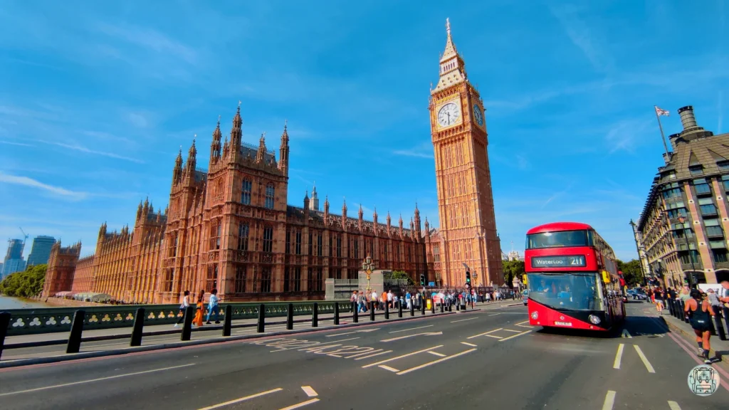 Vista del Big Ben y el Palacio de Westminster en Londres bajo un cielo azul despejado, con un autobús rojo de dos pisos circulando por el puente de Westminster y peatones caminando por la acera. Un destino europeo imprescindible para visitar en julio