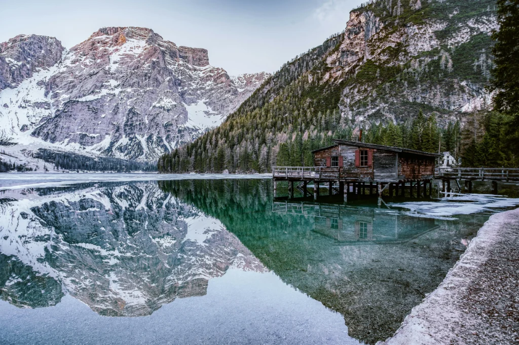 Paisaje alpino del Lago de Braies en Italia al amanecer, con una casa de madera sobre pilotes en el agua cristalina y grandes montañas nevadas reflejándose en la superficie bajo nubes bajas. un destino de naturaleza espectacular donde viajar en junio