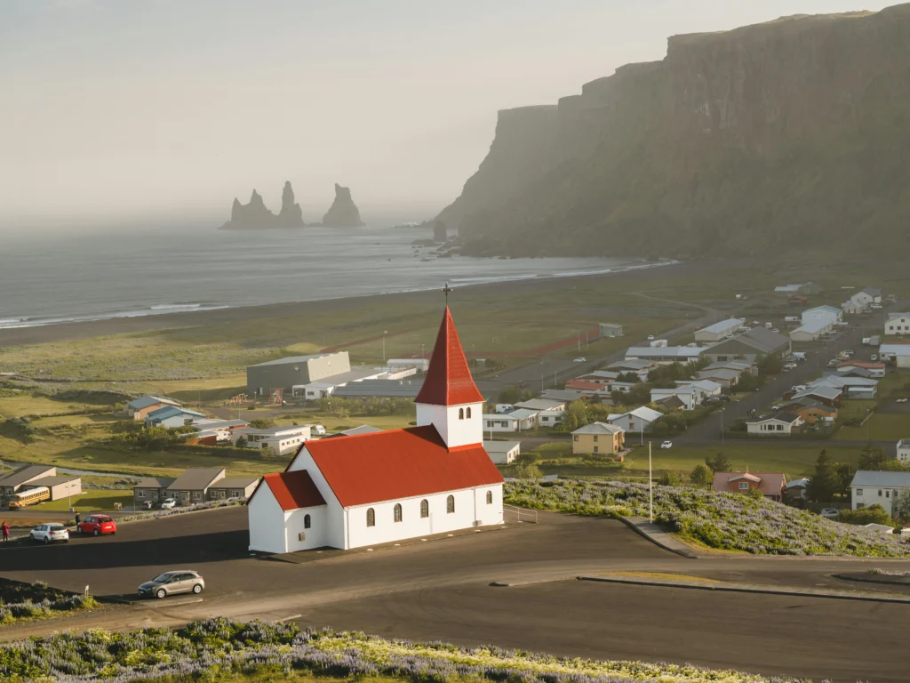 Vista de la iglesia Vik i Myrdal en Islandia bajo un cielo parcialmente nublado, rodeado de prados verdes y el agua azul del fiordo a un lado. ideal para visitar en septiembre