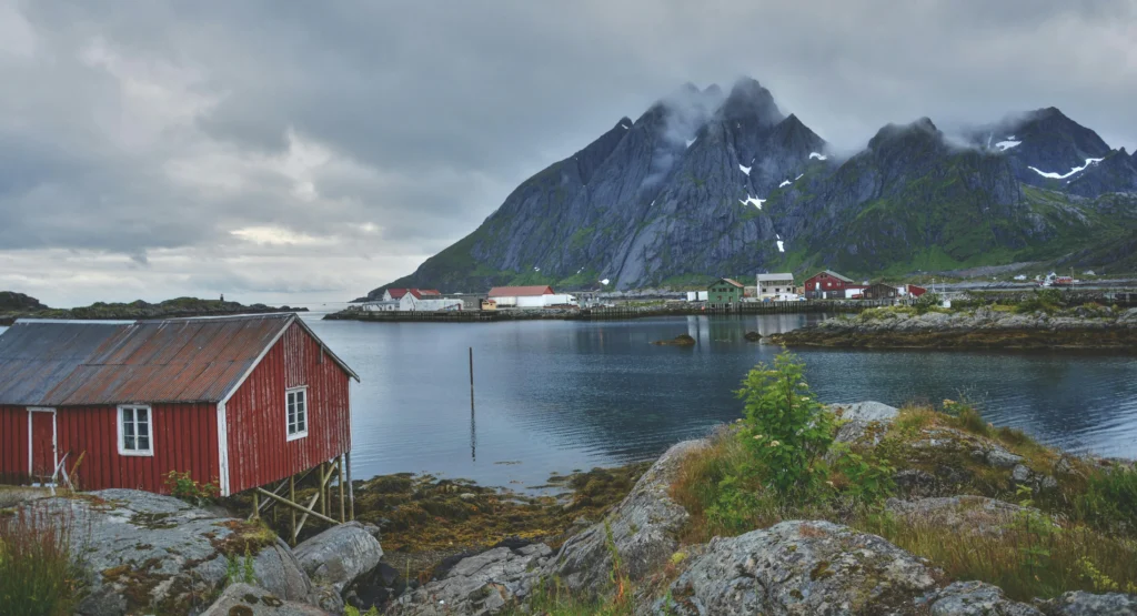 Vista del puerto de Bergen, Noruega, al atardecer, mostrando la hilera de casas históricas de madera de colores iluminadas y reflejadas en el agua, con montañas envueltas en niebla bajo un cielo gris de tormenta.