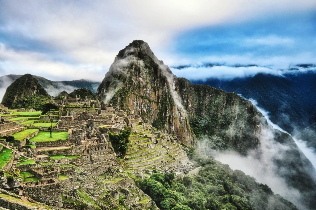 Vista panorámica de la ciudadela inca de Machu Picchu en Perú, con sus terrazas agrícolas verdes y estructuras de piedra rodeadas de nubes bajas y las imponentes montañas de los Andes bajo un cielo iluminado. Destino para viajar en junio